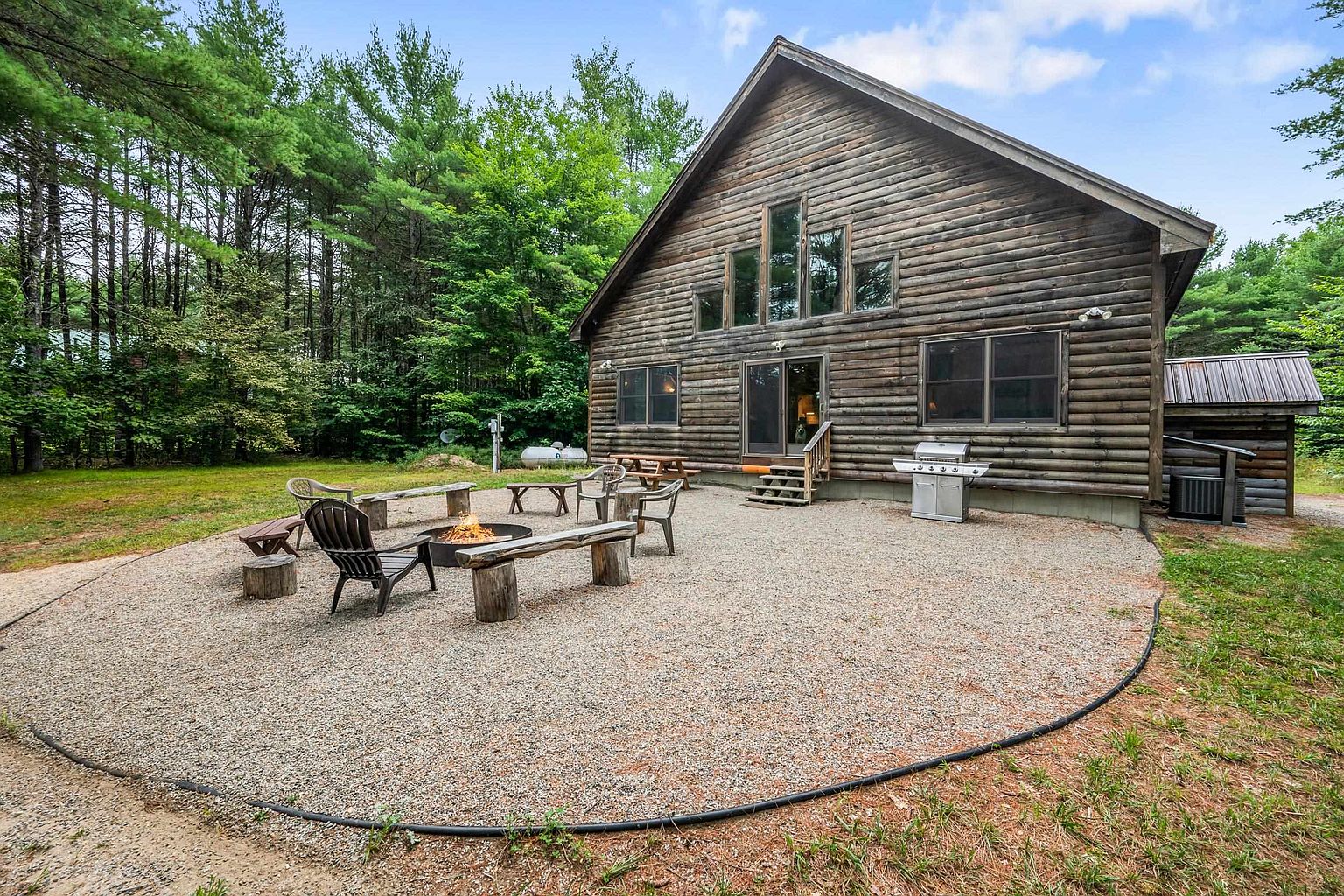 This rear view of a rustic log cabin showcases a spacious gravel patio area complete with a fire pit and outdoor seating. The home features dark wood log construction, a series of tall windows, and a sliding glass door leading to the backyard. The surrounding landscape is heavily wooded, creating a private and serene atmosphere perfect for outdoor relaxation.