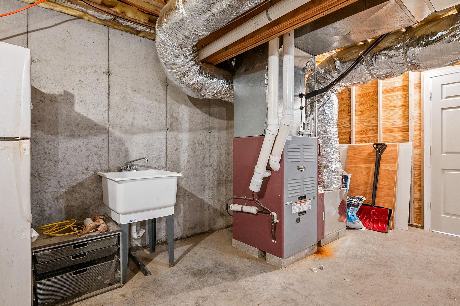 This unfinished basement utility area features exposed concrete walls and a concrete floor, highlighting the mechanical systems of the home. A central furnace unit is connected to overhead silver ductwork, accompanied by a white utility sink and a metal storage unit. The space provides a functional, industrial-style view of the home's infrastructure, including a nearby door and a snow shovel leaning against a plywood-paneled wall.