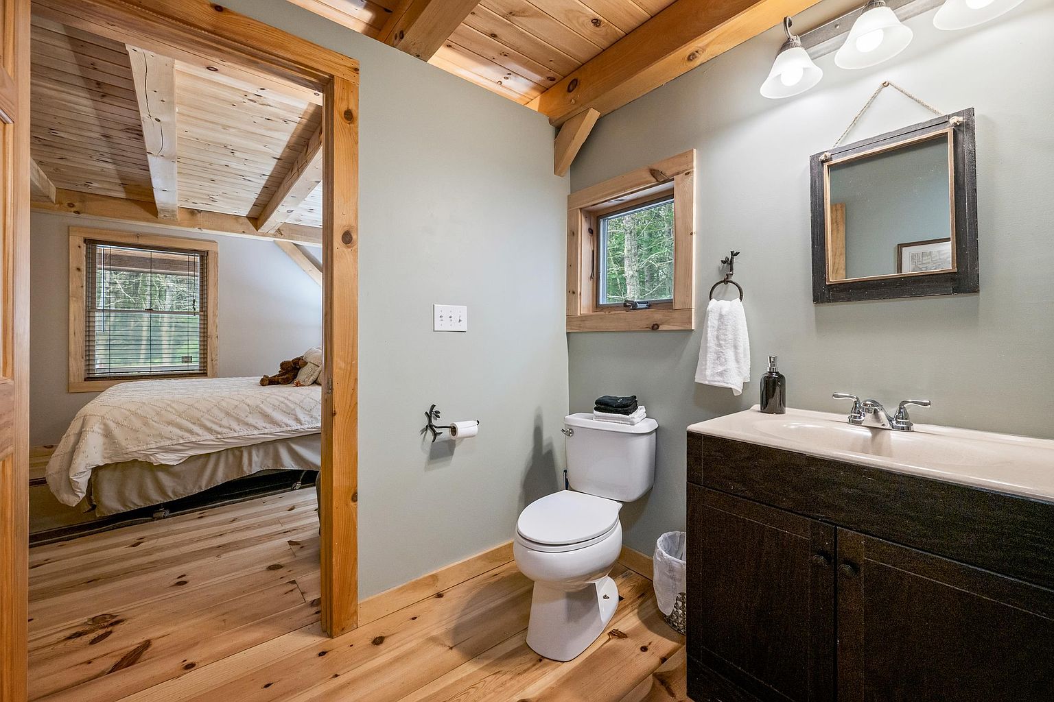This rustic-style bathroom features light-toned wood flooring and a matching wood-paneled ceiling, creating a warm, cabin-like atmosphere. The space includes a white toilet, a dark wood vanity with a light-colored countertop, and a framed mirror hanging above. A doorway provides a direct view into an adjacent bedroom, emphasizing the open and connected layout of the home.