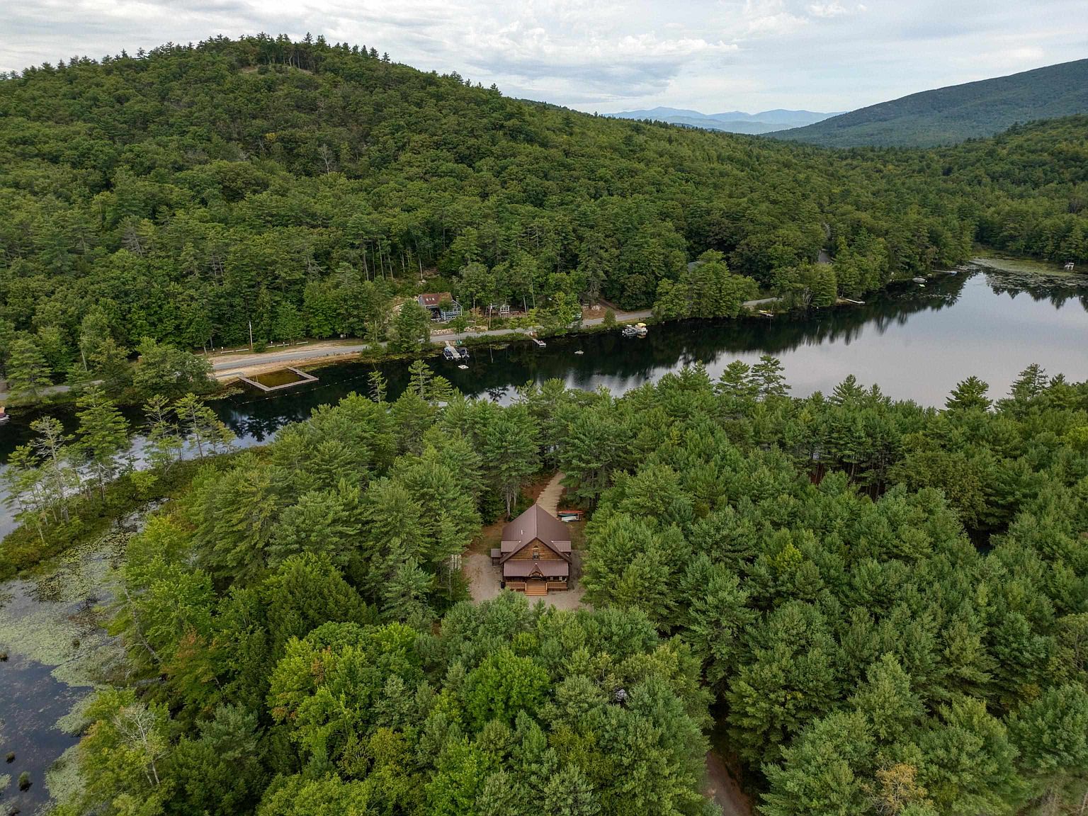 This high-angle aerial shot captures a secluded log-style cabin nestled within a dense, lush forest. The property is situated near a calm, reflective lake, with a road running along the shoreline and rolling green mountains visible in the distance. The perspective emphasizes the home's private, tranquil setting and its integration into the natural landscape.
