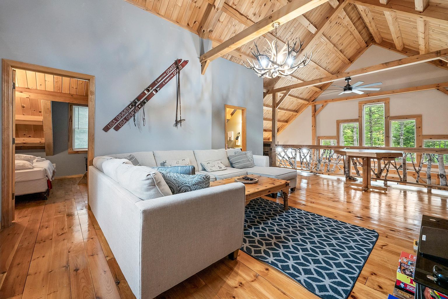 This spacious living room features a vaulted ceiling with exposed wooden beams and a rustic antler chandelier, creating a warm, cabin-like atmosphere. A large, light-colored sectional sofa faces a wooden coffee table, set atop a blue and white patterned area rug on polished wood floors. The room opens to a lofted area with a unique branch-style railing, while vintage skis mounted on the wall add a decorative touch to the light blue accent wall.