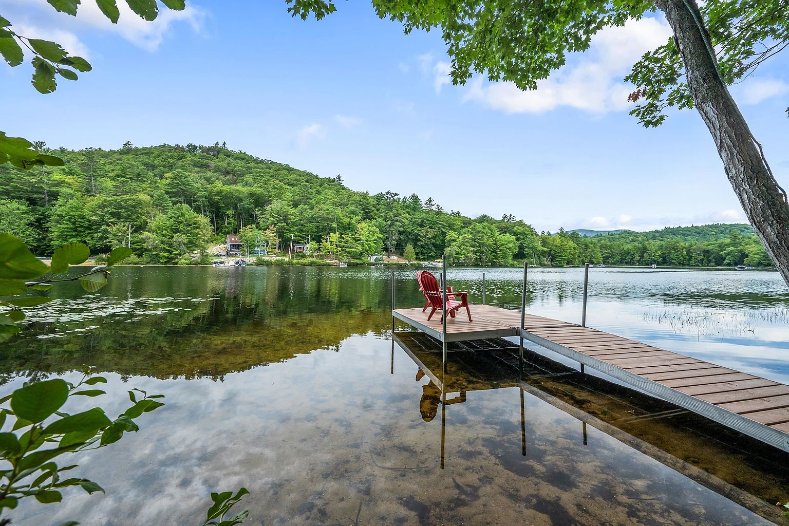 This serene waterfront scene features a wooden dock extending into a calm, clear lake, complete with two red Adirondack chairs for relaxation. The perspective is from the shoreline, looking out across the water toward a lush, green, tree-covered hillside under a bright blue sky with scattered clouds. The tranquil setting highlights the natural beauty and recreational potential of the property's lake access.