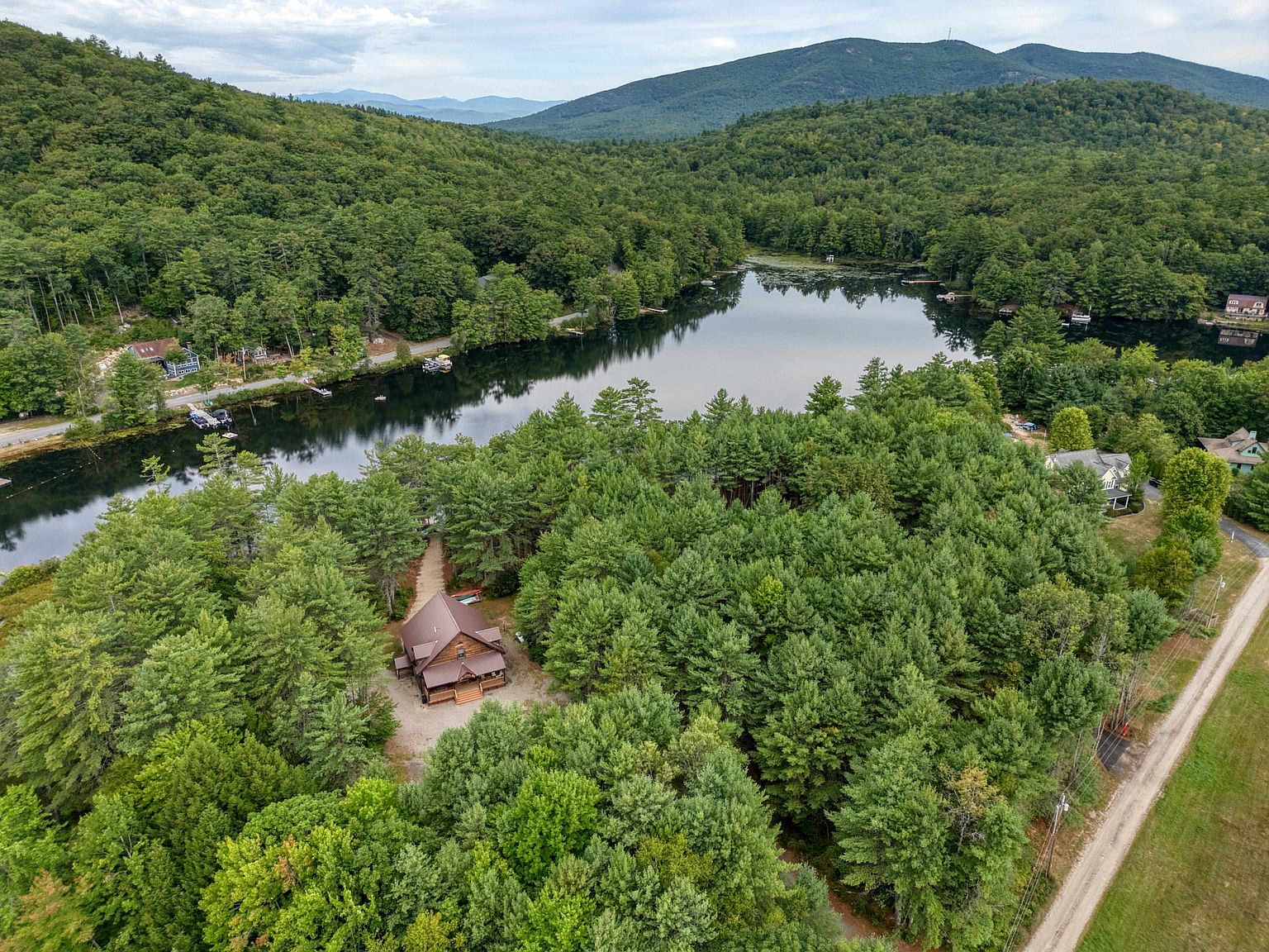 This high-angle aerial perspective captures a secluded log-style cabin nestled within a dense forest, situated near the edge of a serene, reflective lake. The property features a gravel driveway and a private path leading to the water, surrounded by lush greenery and rolling hills in the distance. The scene conveys a peaceful, private retreat atmosphere, highlighting the home's integration with the natural landscape.