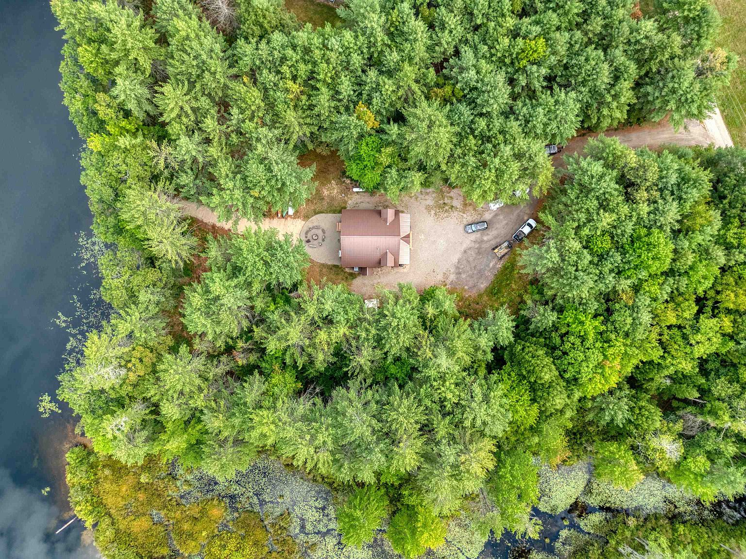 This high-angle aerial view captures a secluded cabin nestled within a dense, lush forest bordering a tranquil lake. The property features a gravel driveway, a small fire pit area, and a metal-roofed structure surrounded by mature trees, offering a private and serene retreat. The perspective emphasizes the natural landscape and the cabin's integration into its wooded environment.