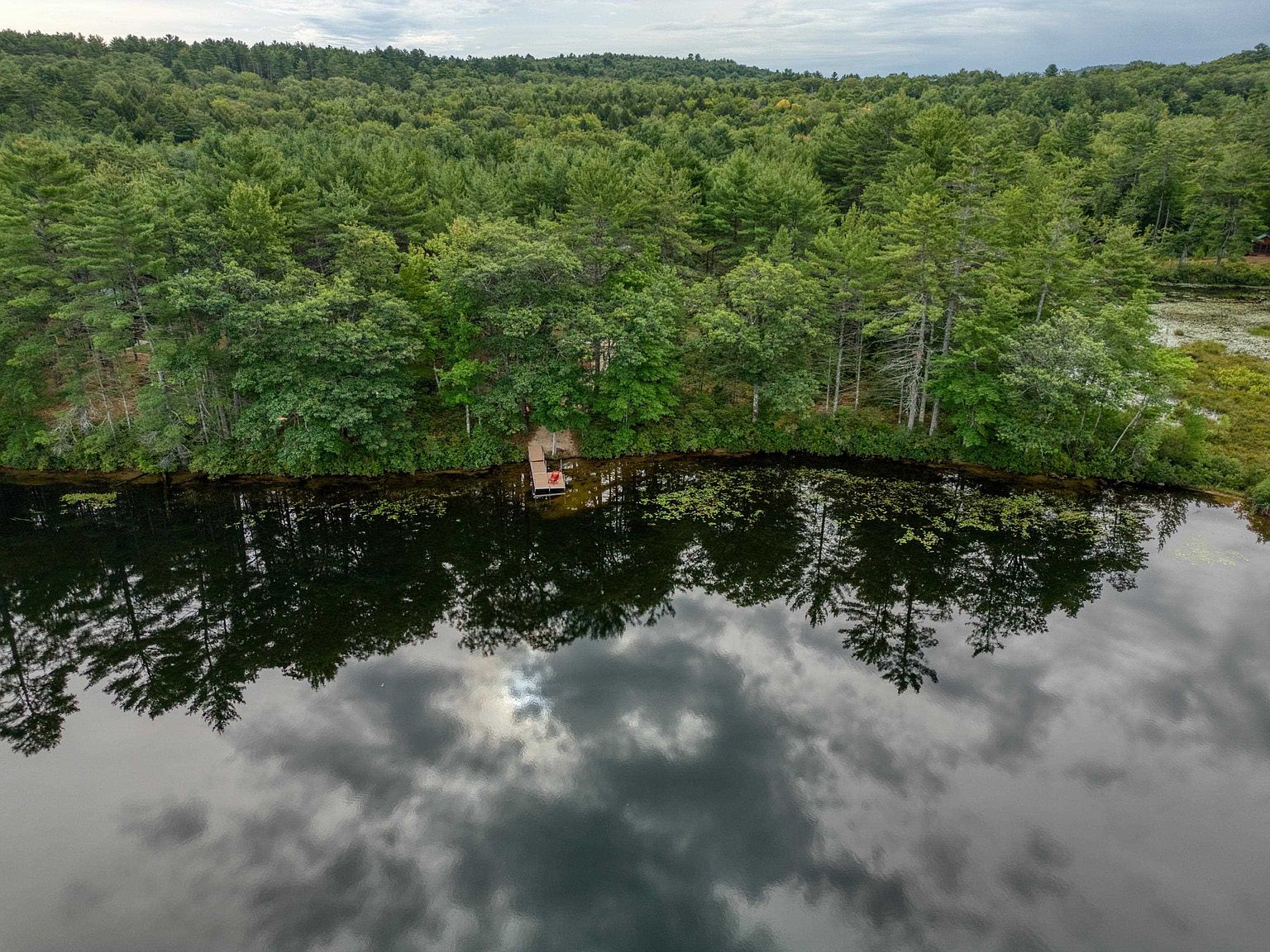This high-angle aerial perspective captures a serene, wooded waterfront property bordering a calm, reflective lake. A small wooden dock extends from the lush, dense forest edge into the water, where a single red chair sits as a focal point. The scene is characterized by the tranquil interplay between the deep green canopy and the mirror-like surface of the lake, which reflects the cloudy sky above.