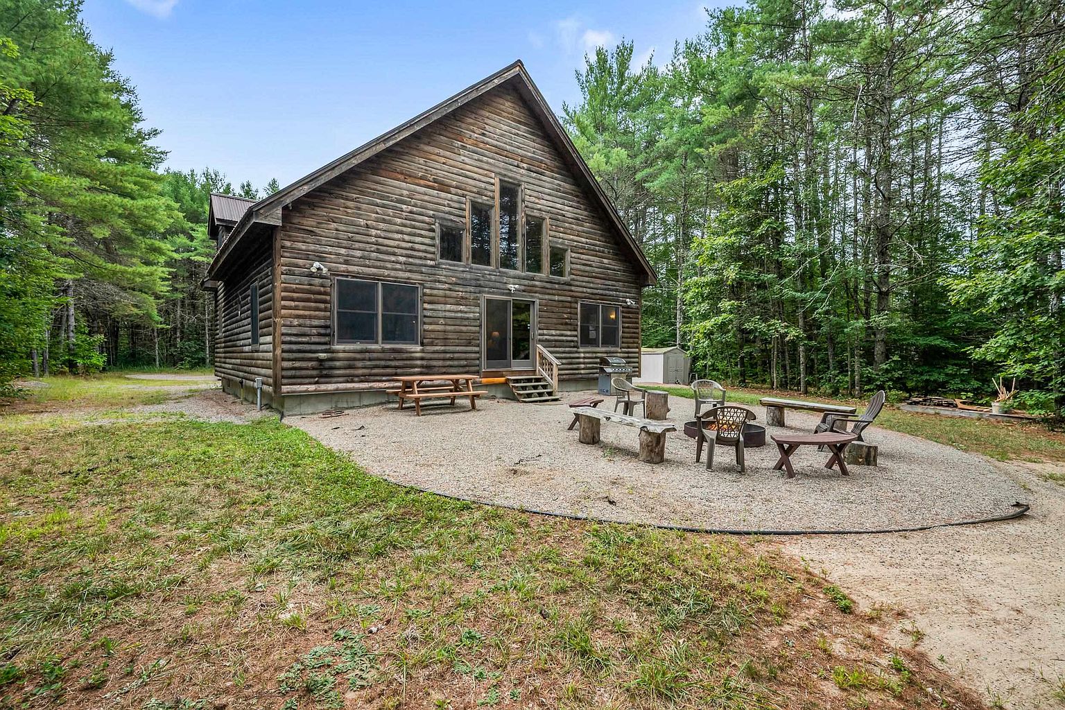 This image captures the rear exterior of a rustic log cabin nestled within a dense, wooded forest. A large gravel patio area features a central fire pit surrounded by various outdoor seating, including wooden benches and chairs, creating an inviting space for relaxation. The cabin itself showcases traditional log construction with a prominent gable roof and a series of tall, vertical windows that allow natural light to filter into the interior.