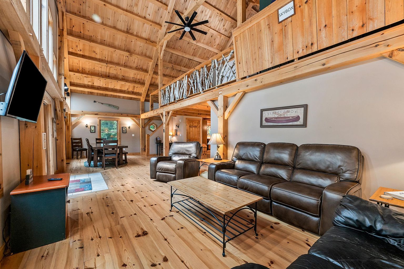 This spacious, open-concept living area features a vaulted ceiling with exposed timber beams and a rustic wood-paneled finish. The room includes a comfortable leather sofa and armchair arrangement, a central coffee table, and a lofted 'skybox' area with a unique birch branch railing. The warm, light-toned wood flooring and natural wood accents create a cozy, lodge-like atmosphere, while the perspective captures both the main seating area and a glimpse of the dining space in the background.