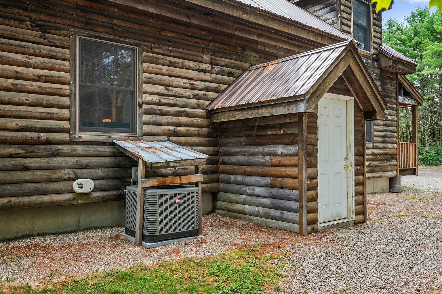 This side view of a rustic log cabin showcases the natural wood exterior and a small, covered entryway with a white door. An outdoor HVAC unit is positioned against the wall, protected by a simple metal-roofed shelter. The ground is covered in gravel, and the surrounding area features mature trees, contributing to a secluded, woodland atmosphere.