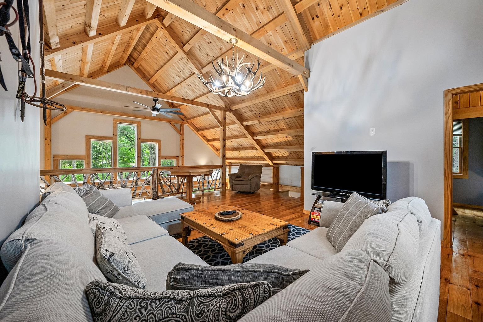 This spacious, rustic-style living room features a vaulted ceiling with exposed timber beams and a distinctive antler chandelier. The room is furnished with a large, light-colored sectional sofa, a wooden coffee table, and a television, all set against a backdrop of warm wood paneling and a lofted area with a railing. Large windows provide natural light and views of the surrounding greenery, creating an inviting and cozy atmosphere.