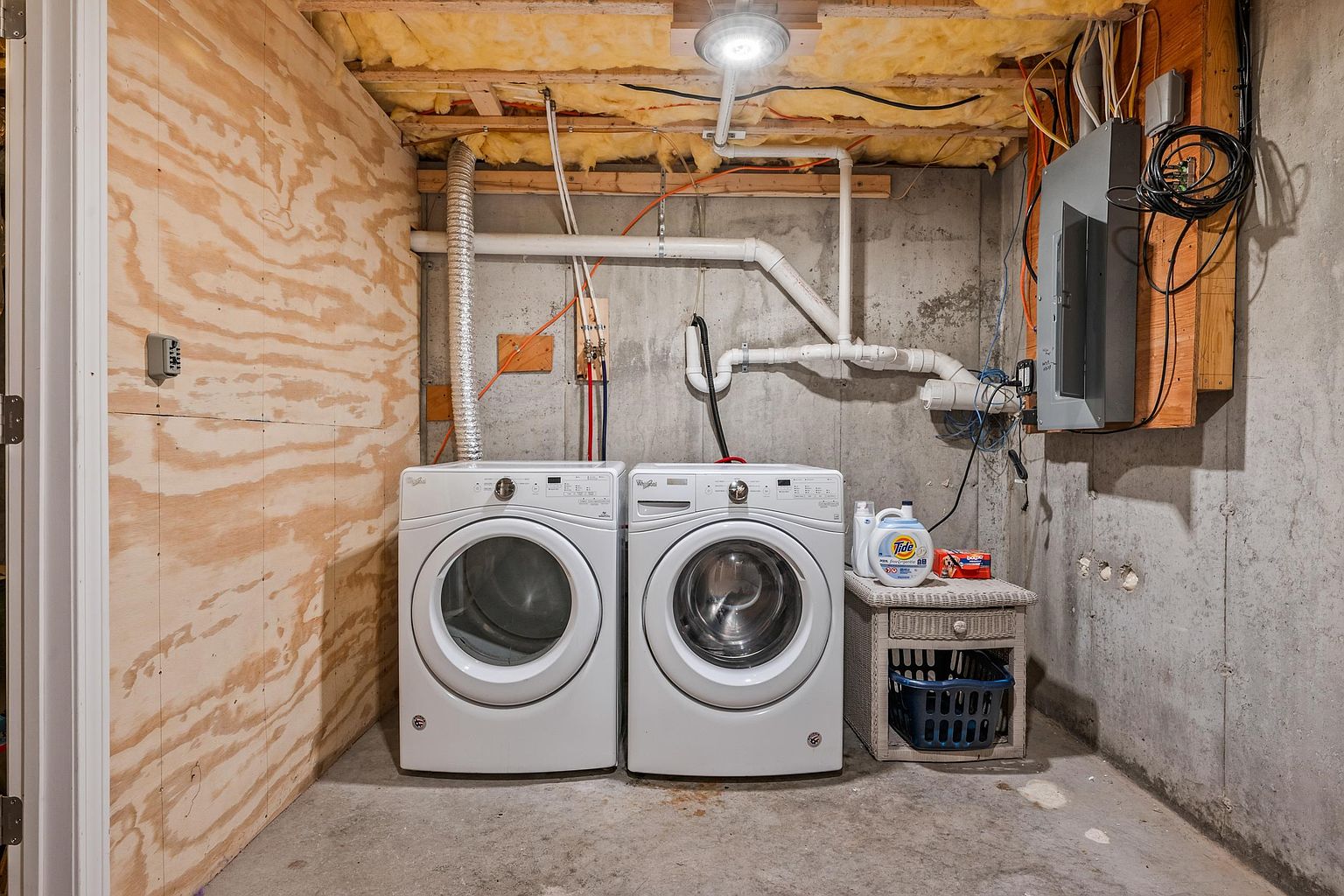 This unfinished basement laundry area features a pair of white front-loading Whirlpool appliances set against a concrete foundation wall. To the left, a plywood-paneled wall adds a rustic texture, while the ceiling remains exposed with visible insulation and wiring. A small wicker side table holds laundry detergent, providing a functional and utilitarian space.