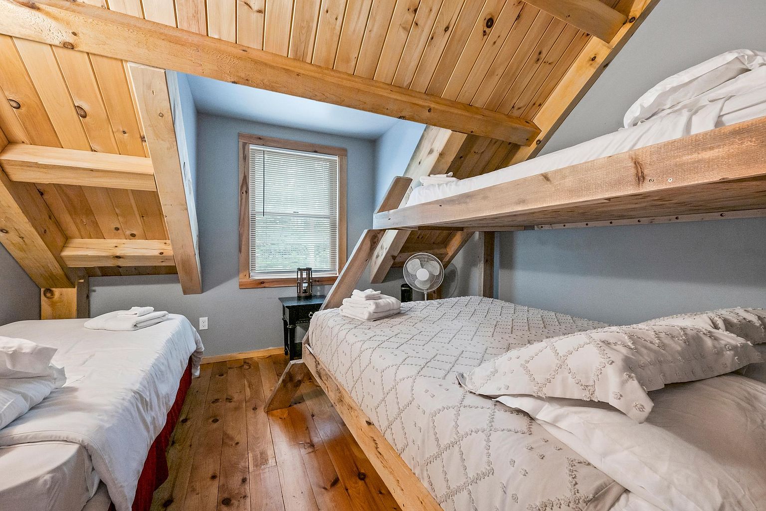 This cozy bedroom features a rustic aesthetic with exposed wood beams on the vaulted ceiling and warm wood flooring. The room is furnished with a bunk bed and an additional single bed, both dressed in white linens, making it an efficient space for multiple guests. A small window with blinds allows natural light into the room, which is accented by a small black side table and a portable fan.