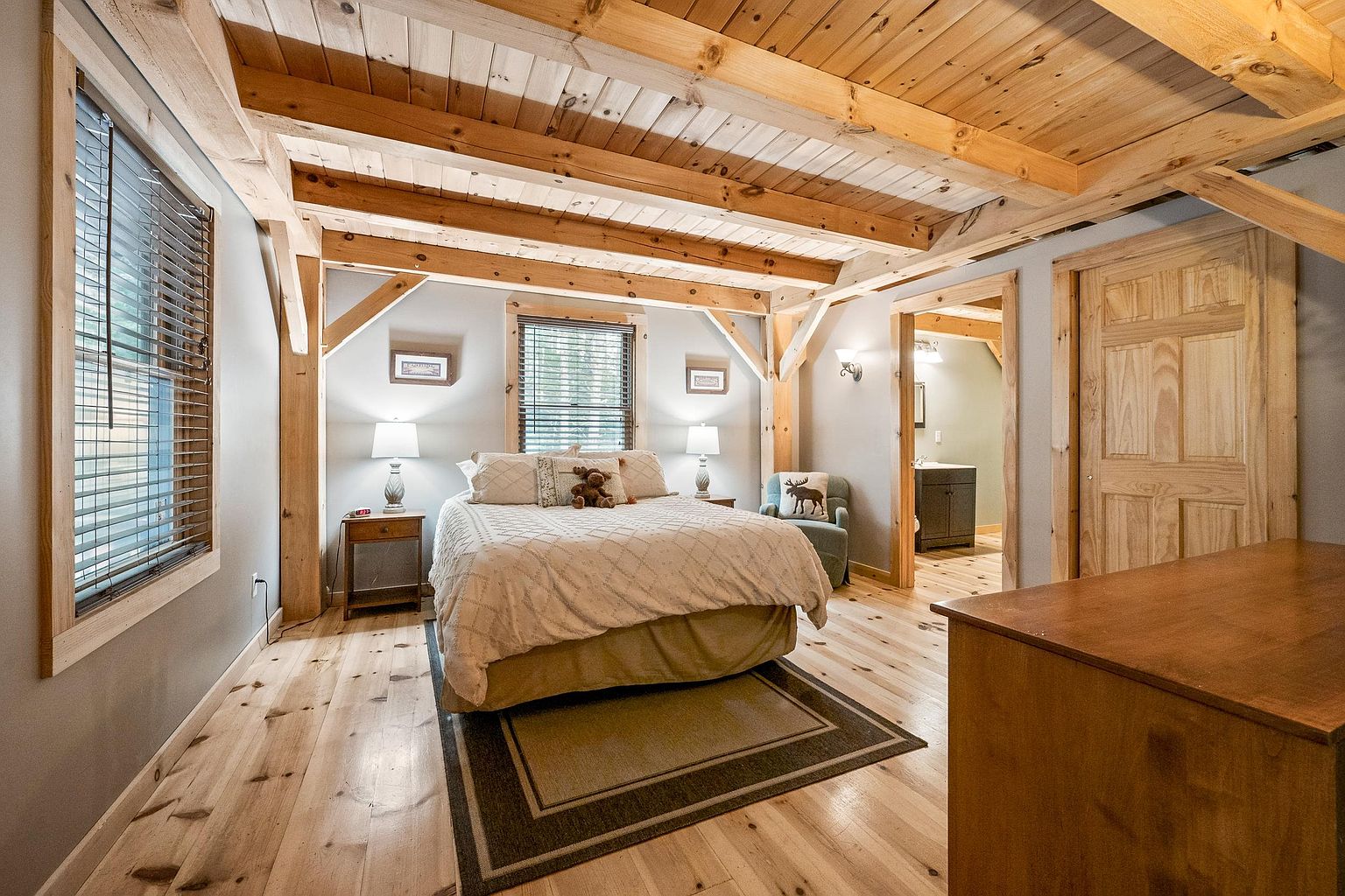 This rustic-style bedroom features exposed timber beams on the ceiling and light wood flooring, creating a warm and inviting atmosphere. The room is centered around a queen-sized bed with a neutral-toned quilt, flanked by two matching nightstands and lamps. A doorway leads into an adjacent bathroom, while a large window on the left allows natural light to fill the space, highlighting the cozy, cabin-inspired aesthetic.