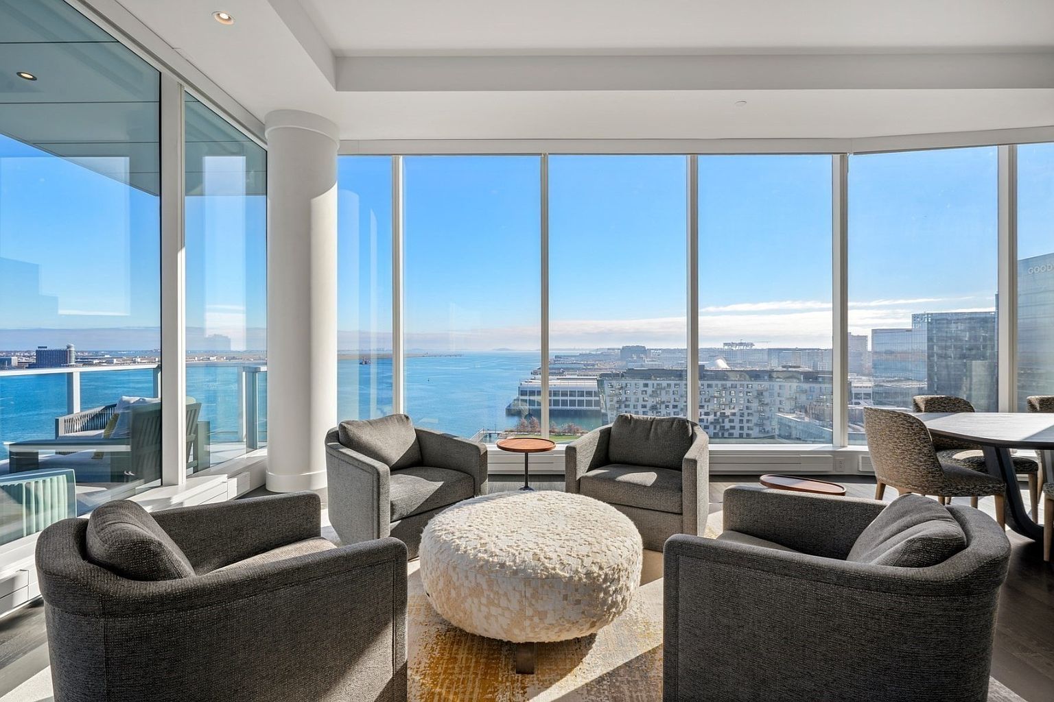 This is a bright and modern living room with floor-to-ceiling windows offering panoramic views of the water and city skyline. The room features a seating arrangement with four gray armchairs surrounding a round, textured ottoman on a patterned rug. A dining table with chairs is visible in the background, and the overall aesthetic is luxurious and contemporary.