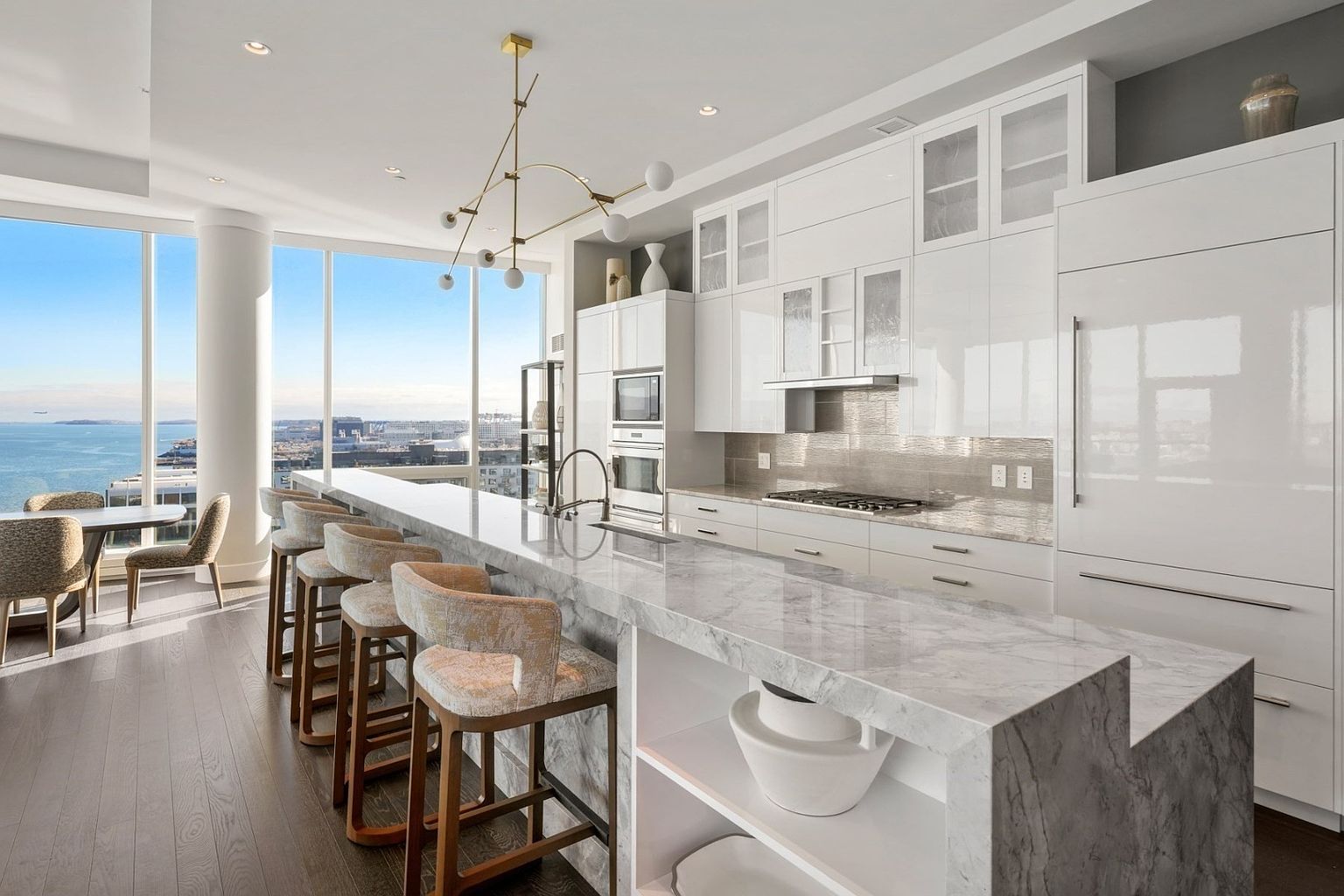 This is a bright and modern kitchen featuring sleek white cabinetry, stainless steel appliances, and a large marble island with seating. The kitchen boasts a stunning view through large windows, enhancing the open and airy feel. A stylish chandelier hangs above the island, adding a touch of elegance.