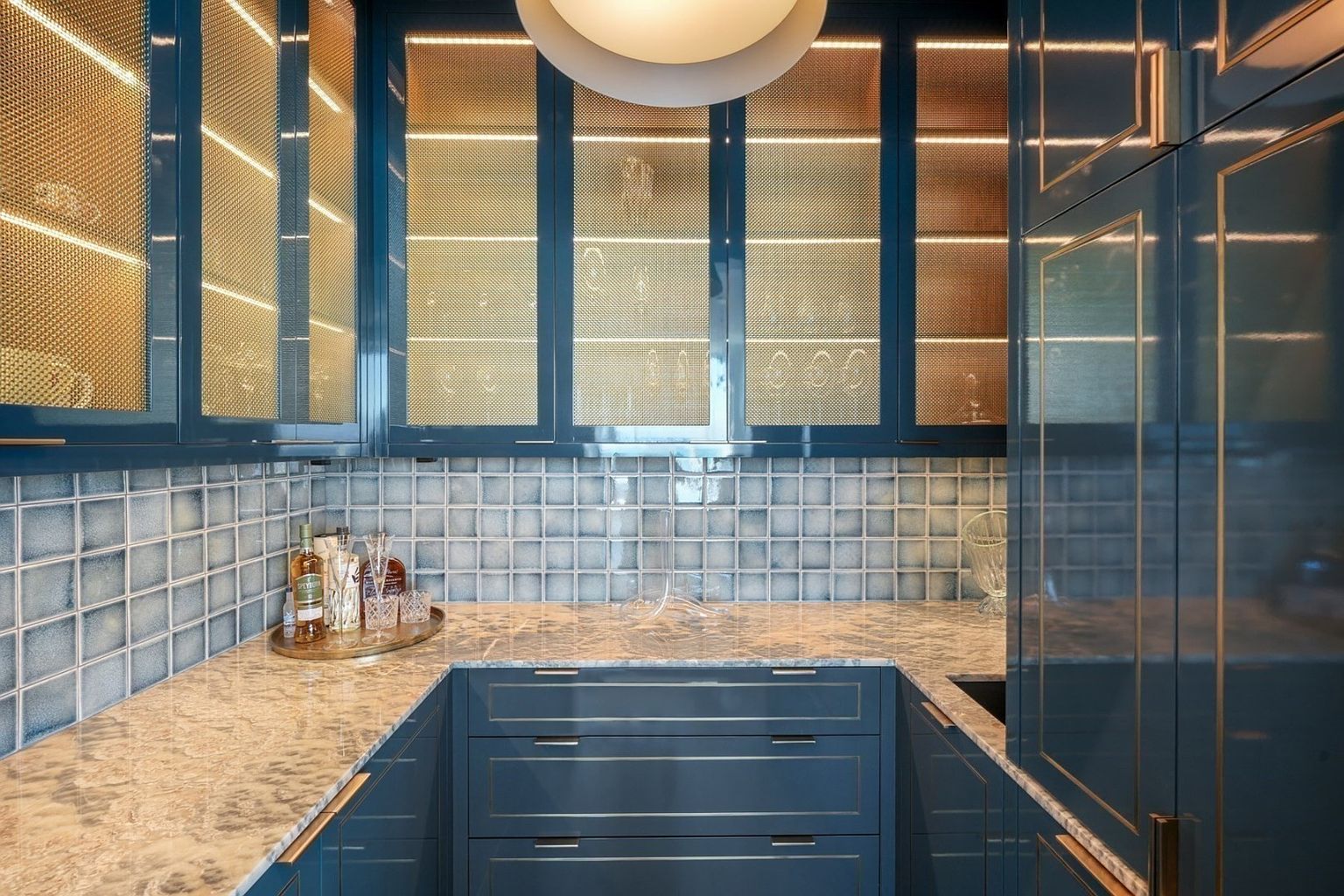 This image showcases a stylish kitchen area with custom blue cabinetry accented by gold trim. The upper cabinets feature a unique mesh front, illuminated from within, displaying glassware. The countertop is a light marble, and the backsplash is composed of small, textured tiles, creating a sophisticated and modern aesthetic.