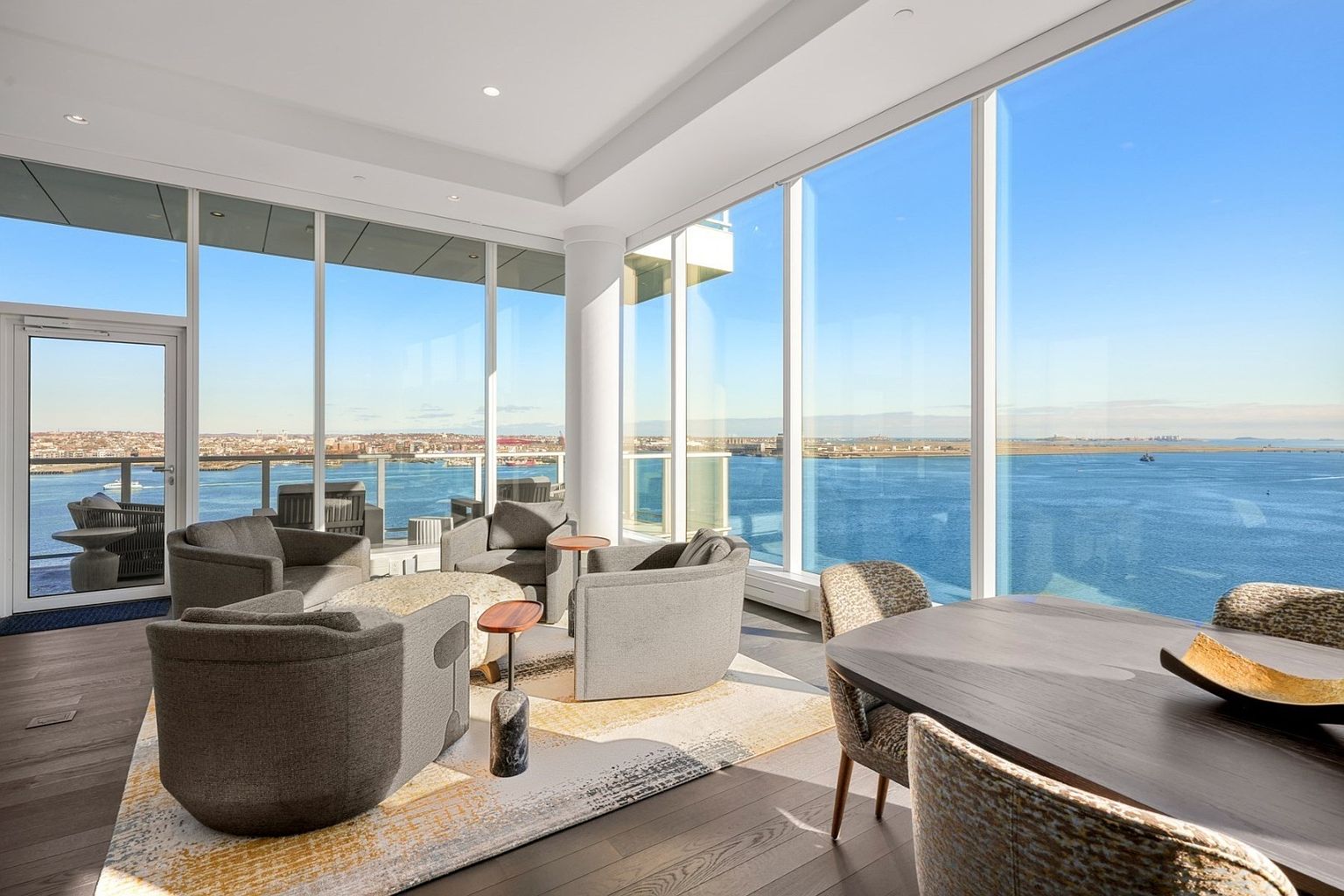 This is a bright and airy living room with floor-to-ceiling windows offering panoramic water views. The room features a modern design with comfortable gray armchairs arranged around a central rug and a dark wood dining table with upholstered chairs. The space is well-lit and inviting, perfect for relaxation and entertaining.
