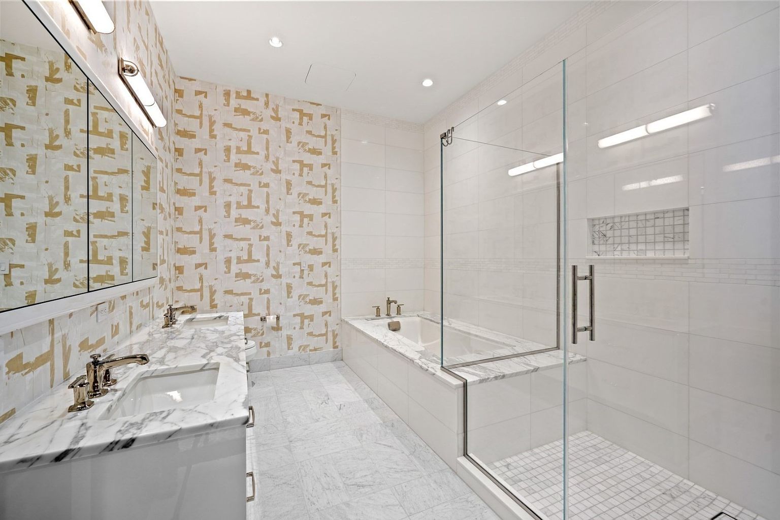 This is a luxurious primary bathroom featuring a double vanity with marble countertops, a large mirror, and patterned wallpaper. The bathroom also includes a separate shower and bathtub enclosed by glass, with white tile throughout. The overall impression is clean, modern, and upscale.