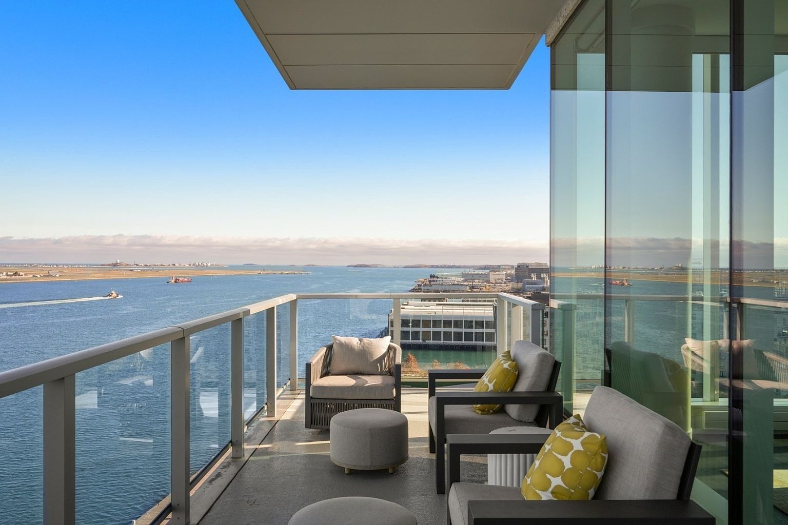 This image showcases a luxurious balcony with comfortable outdoor seating, including armchairs and ottomans, set against a stunning ocean view. The balcony features a modern glass railing, providing an unobstructed panorama of the water and distant cityscape. The overall impression is one of upscale relaxation and sophisticated outdoor living.