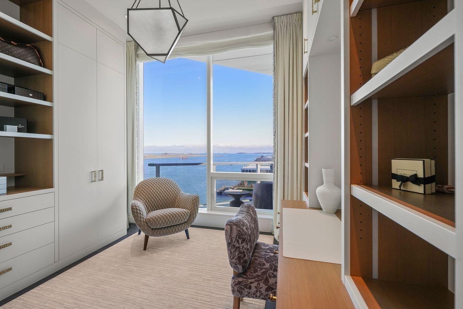 This is a well-organized walk-in closet with custom shelving and cabinetry. A seating area with a patterned chair and a view of the ocean adds a luxurious touch. The space is bright and airy, making it an appealing feature for potential buyers.