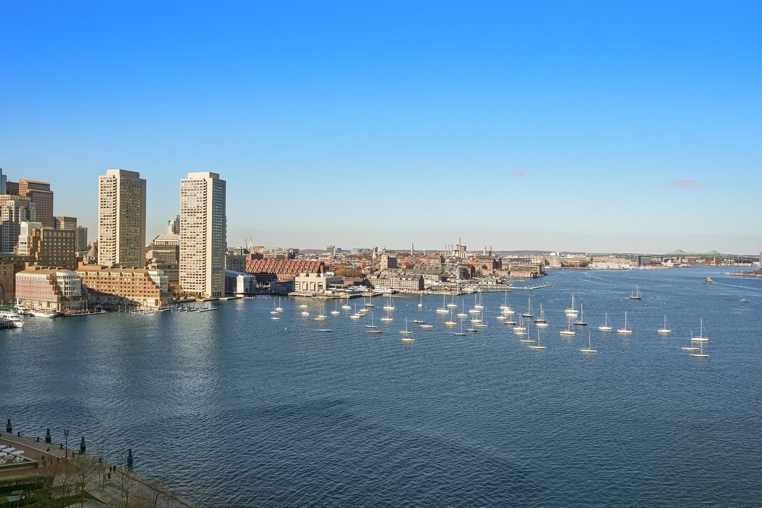 This aerial view showcases a stunning cityscape with modern high-rise buildings along the waterfront. Numerous sailboats dot the expansive blue harbor, creating a picturesque scene. The clear blue sky enhances the overall appeal, suggesting a vibrant and desirable urban environment.