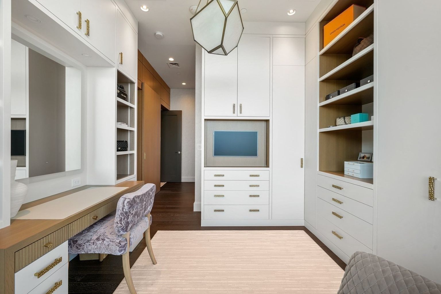 This is a well-organized walk-in closet featuring custom white cabinetry with gold hardware, open shelving, and a built-in vanity area. The closet includes a television, drawers, and ample storage space, creating a luxurious and functional dressing area. The dark wood flooring contrasts with the light-colored rug, adding depth to the space.
