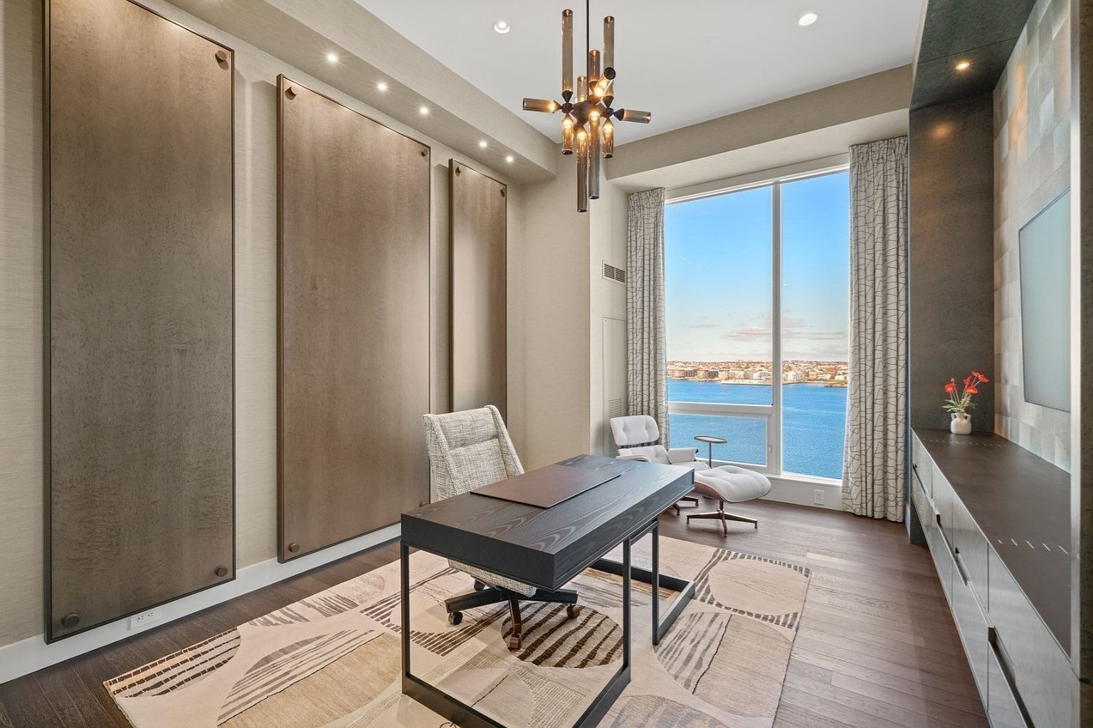 This is a modern office space featuring a dark wood desk with a light-colored chair, complemented by a contemporary rug with geometric patterns. The room is well-lit with a unique chandelier and recessed lighting, and large windows offer a stunning view of the water. The overall design is sophisticated and functional, creating an ideal workspace.