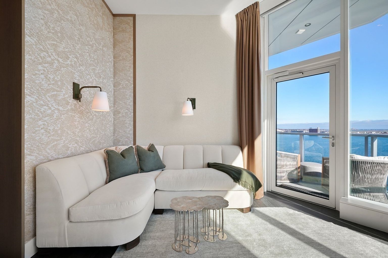 This is an interior shot of a living room featuring a curved, off-white sectional sofa with green pillows and a throw blanket. A unique, multi-tiered side table sits beside the sofa, and a large window offers a stunning view of the ocean and cityscape. The room is decorated in neutral tones with textured wallpaper, creating a serene and luxurious atmosphere.