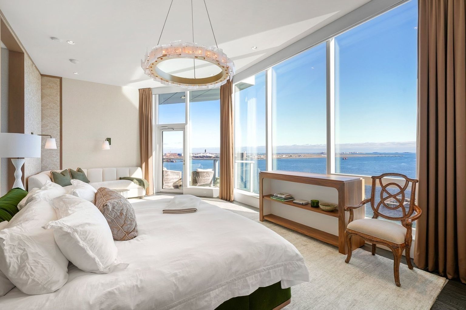 This is a luxurious primary bedroom with floor-to-ceiling windows offering stunning ocean views. The room features a large bed with white linens, a seating area, and a modern chandelier. A wooden console table and an elegant chair add to the sophisticated ambiance, creating a serene and upscale retreat.