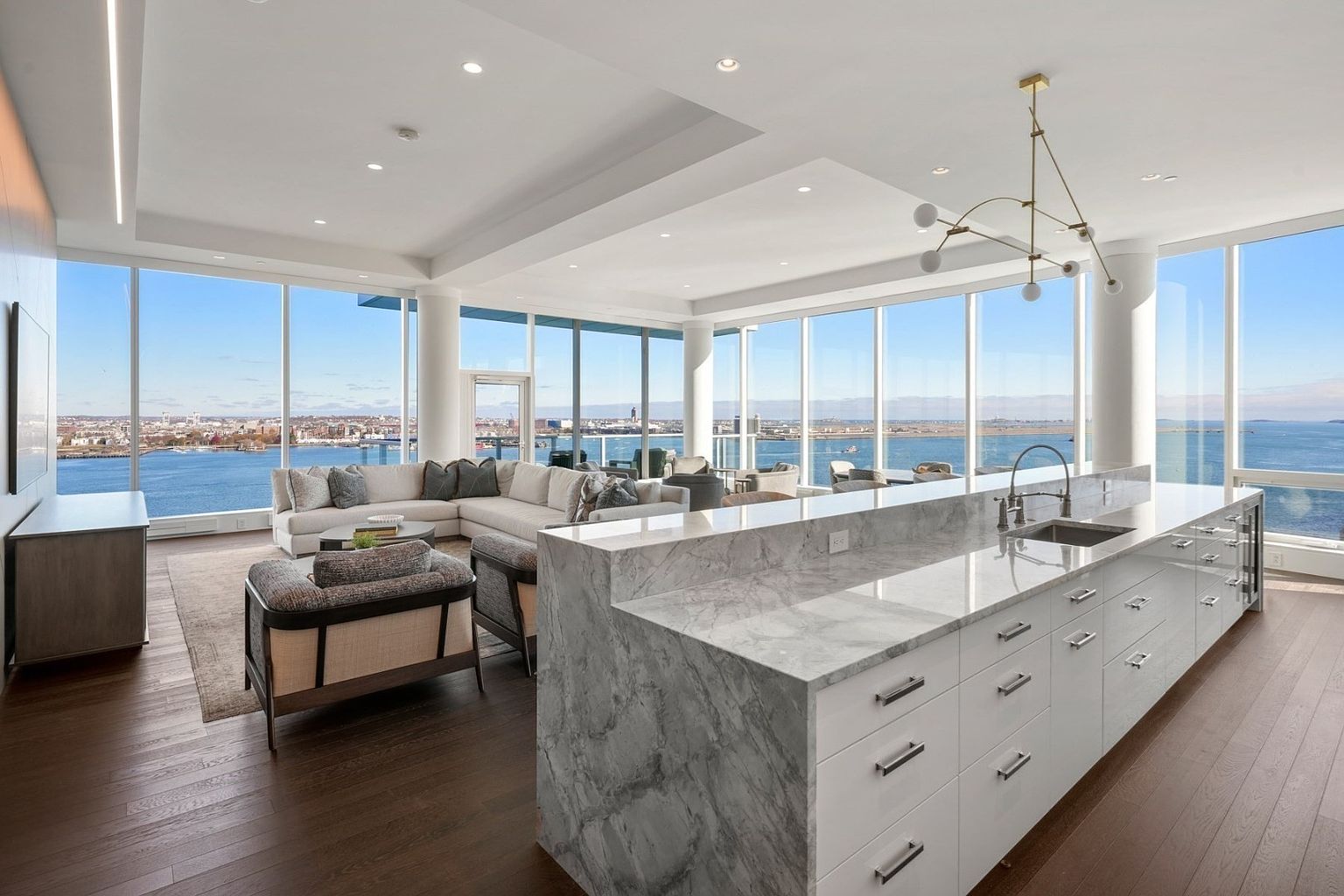This is a bright and modern kitchen and living area with floor-to-ceiling windows offering panoramic water views. The kitchen features a large island with marble countertops and white cabinetry, complemented by a stylish chandelier. The living area includes a comfortable sectional sofa and contemporary seating, creating an open and inviting space.