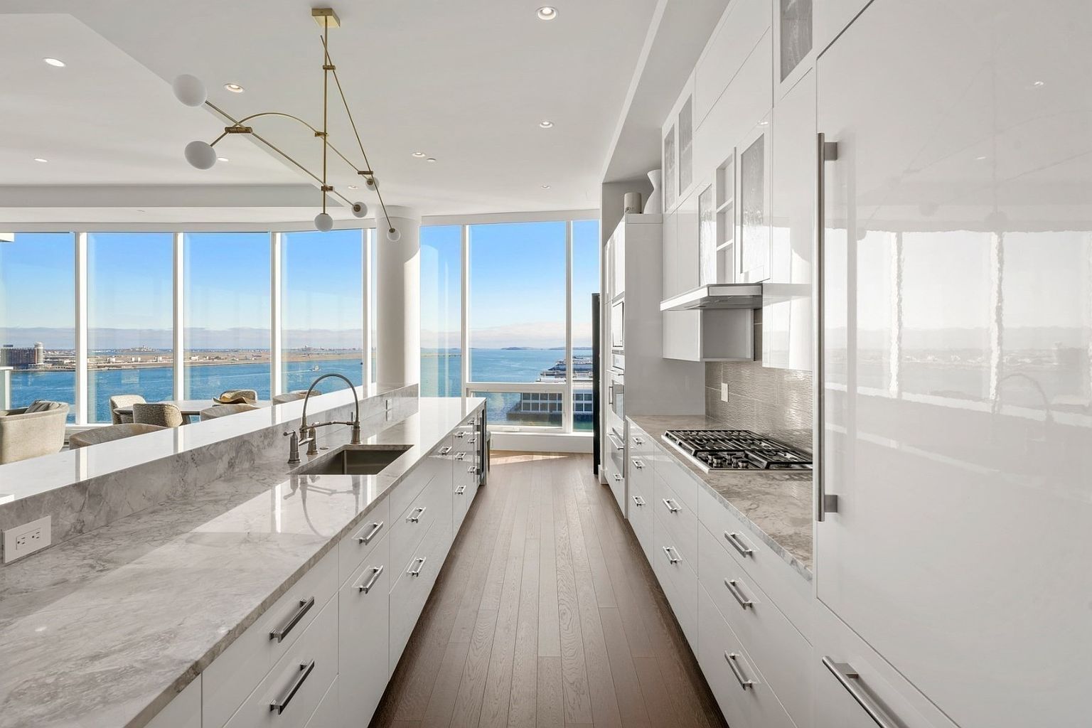 This is a bright, modern kitchen with sleek white cabinetry and countertops. Large windows offer a stunning view of the water and cityscape. The kitchen features stainless steel appliances and a contemporary lighting fixture, creating a luxurious and inviting space.