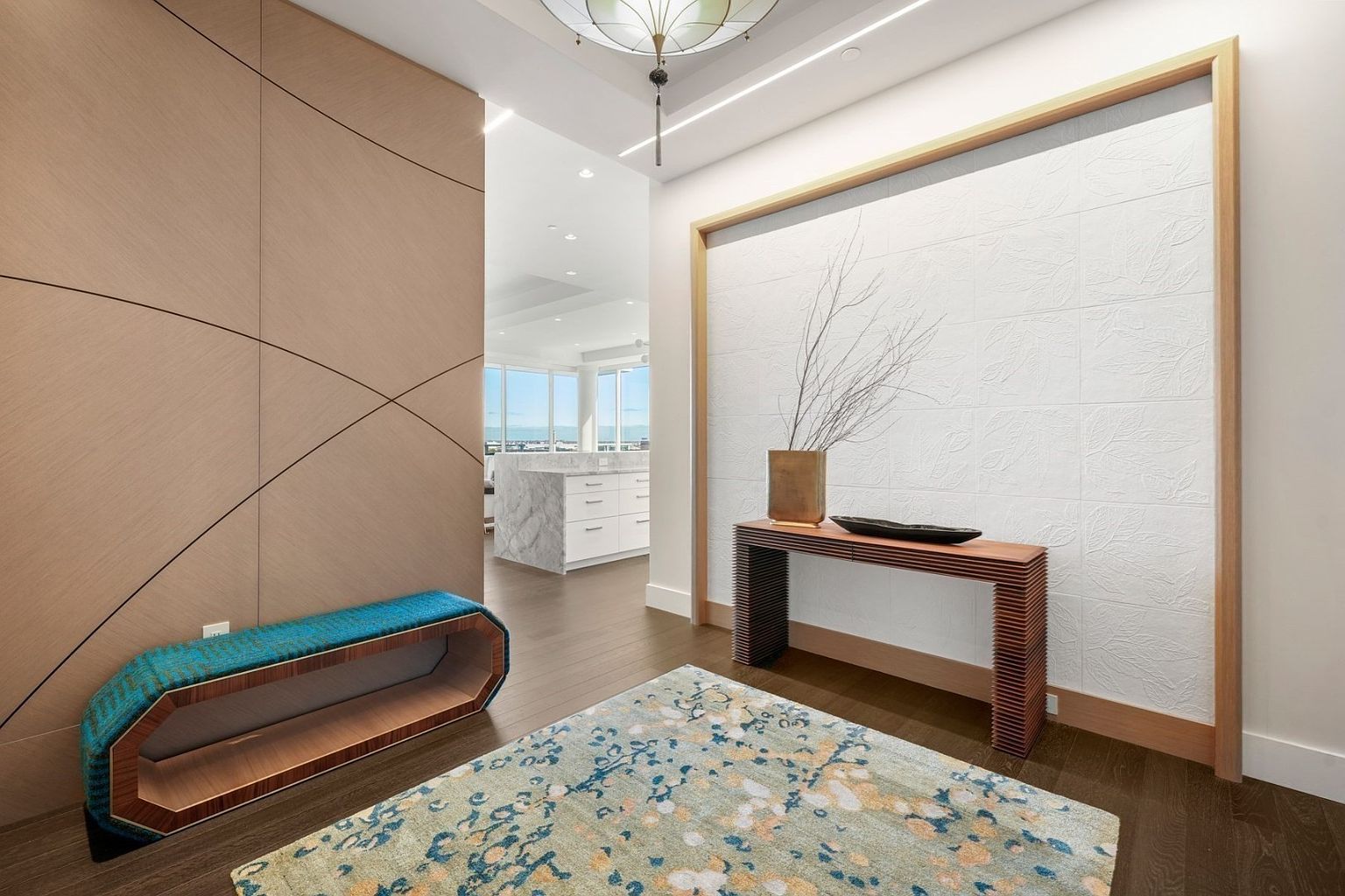 This interior shot showcases a modern hallway with a unique blend of textures and design elements. A textured wooden wall panel complements a blue upholstered bench, while a decorative rug adds a splash of color to the dark wood flooring. The hallway leads into a bright, open kitchen area, creating a sense of flow and spaciousness.