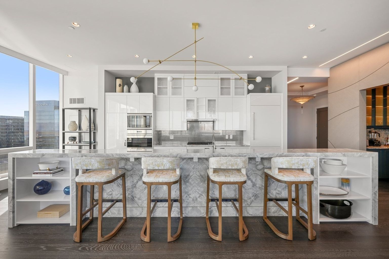 This is a bright and modern kitchen featuring a large marble island with seating for four. The kitchen is equipped with white cabinetry, stainless steel appliances, and a unique gold chandelier. Large windows provide natural light and views of the city.
