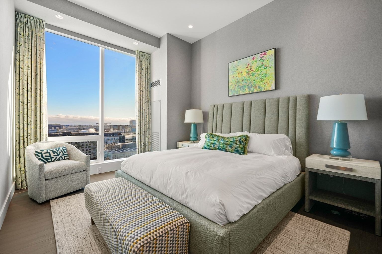 This is a primary bedroom featuring a large bed with a padded headboard, white linens, and a decorative pillow. The room has a neutral color palette with gray walls and a large window offering a city view. A patterned bench sits at the foot of the bed, and there is a comfortable armchair in the corner, creating a relaxing and luxurious atmosphere.