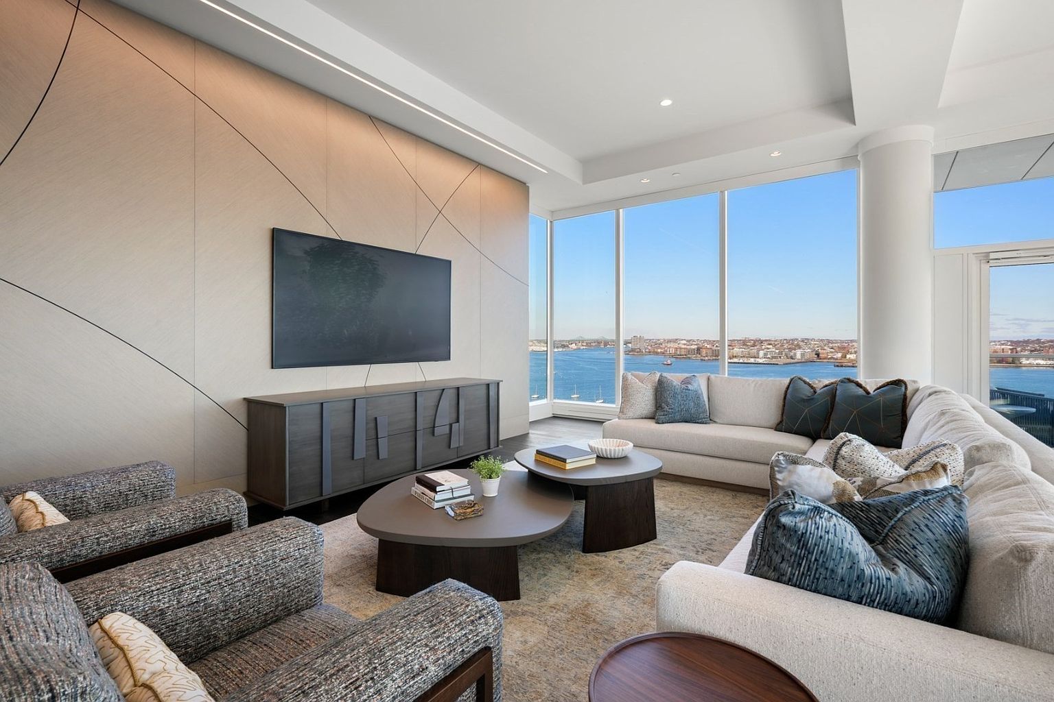 This is a modern living room with a large sectional sofa, two armchairs, and a media console with a large television. The room features floor-to-ceiling windows with a view of the water and city skyline. The color palette is neutral with pops of blue, creating a sophisticated and inviting space.