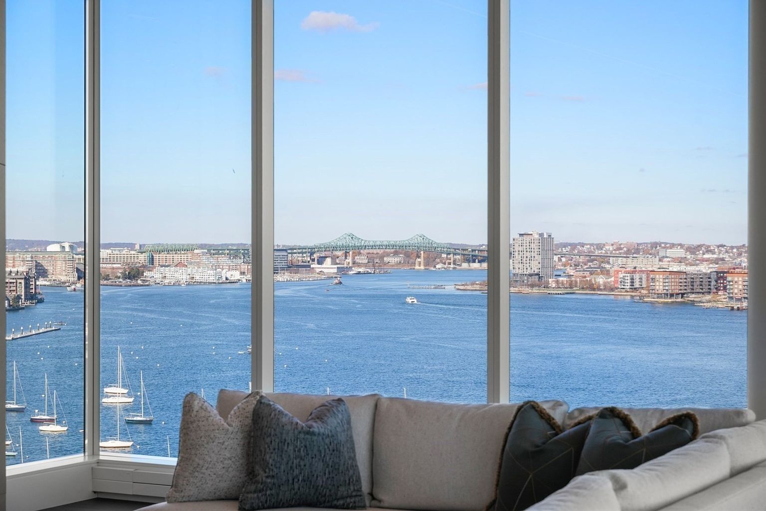 This image showcases a luxurious living room with a stunning panoramic view of a waterfront city skyline, visible through expansive floor-to-ceiling windows. A comfortable, neutral-toned sofa with decorative pillows sits in the foreground, inviting relaxation and enjoyment of the breathtaking scenery. The overall impression is one of sophisticated urban living with a focus on the spectacular view.