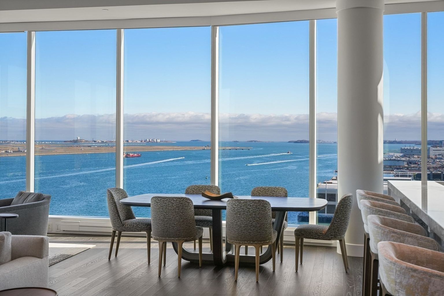 This is a bright and airy dining room with floor-to-ceiling windows offering panoramic water views. The room features a modern dining table with six chairs, complemented by hardwood flooring and a neutral color palette. A nearby bar area with stools adds to the luxurious feel of the space.