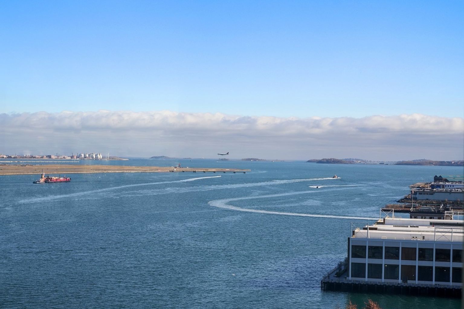 This aerial view showcases a waterfront property with a modern building featuring large windows and a flat roof. The scene includes a vast expanse of water with boats leaving wakes, a distant shoreline, and an airplane in the sky. The overall impression is one of spaciousness and a prime waterfront location.