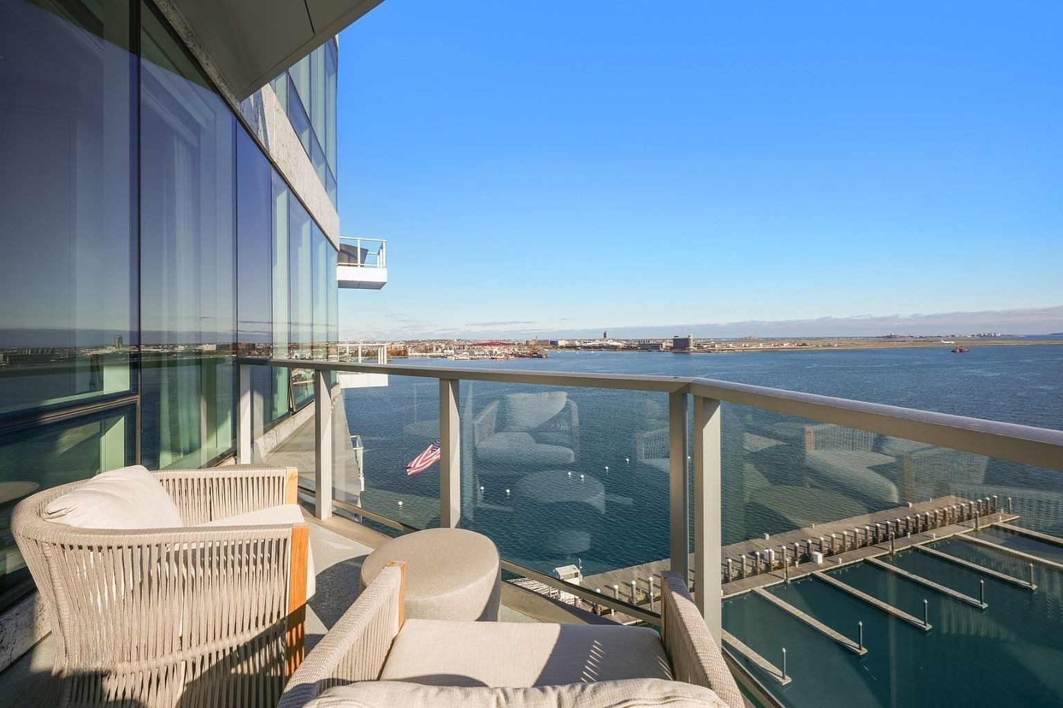 This image showcases a luxurious balcony with comfortable outdoor seating, offering stunning waterfront views. The balcony features modern glass railings and overlooks a marina with boat slips. The scene evokes a sense of upscale living and relaxation with a clear blue sky in the background.