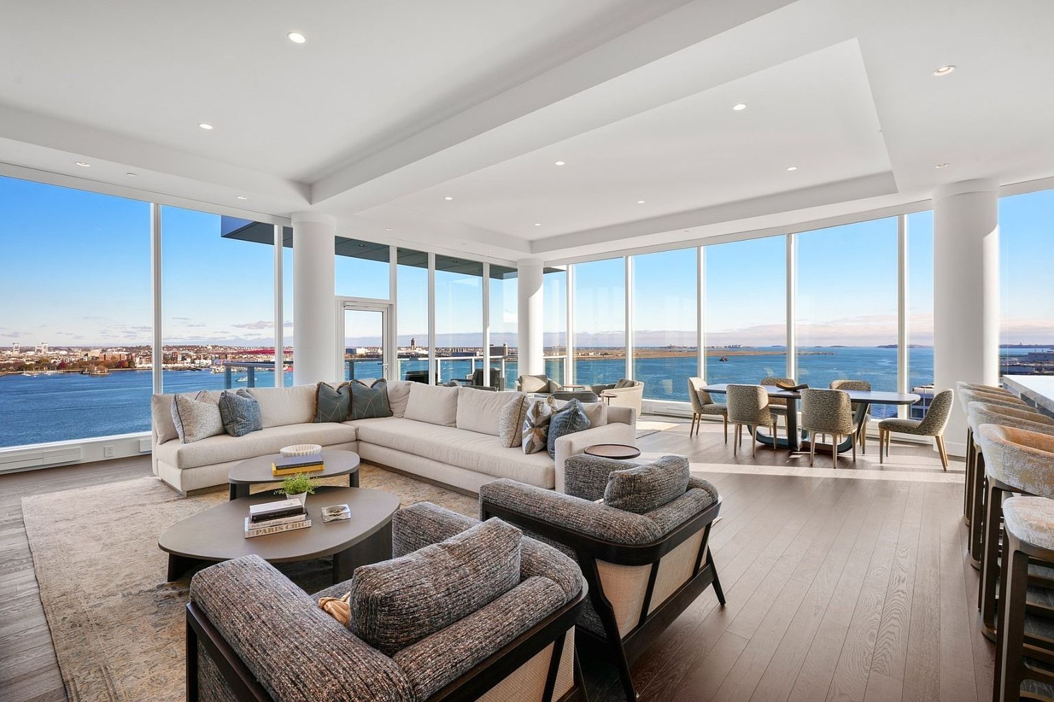 This is a spacious and luxurious living room with floor-to-ceiling windows offering panoramic water views. The room features a large sectional sofa, two armchairs, and a round coffee table, creating a comfortable and inviting atmosphere. The hardwood floors and neutral color palette contribute to the room's sophisticated and modern aesthetic.