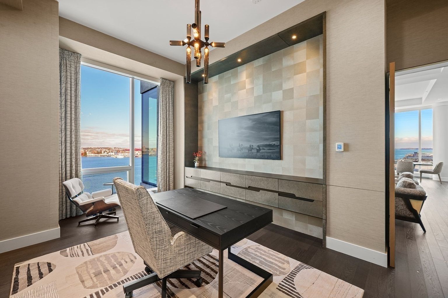 This is a well-appointed home office featuring a modern desk and chair, a large window with a water view, and a built-in media center with a television. The room is decorated in neutral tones with a patterned rug, creating a sophisticated and comfortable workspace. The perspective is from the doorway, showcasing the entire office space.