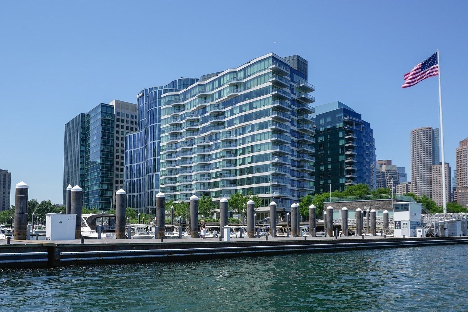 This image showcases a modern waterfront property with a striking architectural design. The building features multiple stories with glass balconies, offering stunning views. A pier extends into the water, adding to the property's appeal and providing direct access to the waterfront. An American flag waves in the background.