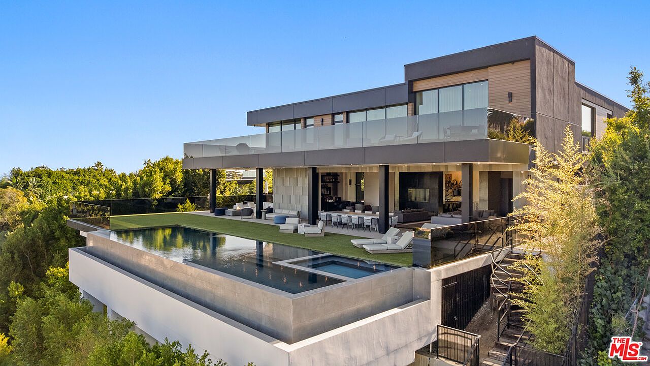 This image showcases the rear exterior of a modern luxury home, emphasizing its multi-level design and outdoor living spaces. A large infinity pool and spa are prominent features, complemented by a manicured lawn and multiple seating areas. The architecture is characterized by clean lines, glass railings, and a seamless integration with the surrounding lush greenery, creating an inviting and sophisticated atmosphere.