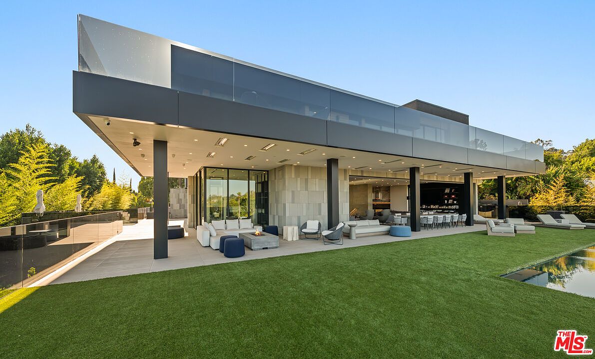 This image showcases the rear exterior of a modern luxury home, featuring a sleek, cantilevered design with extensive glass walls and a spacious covered patio. The outdoor area includes a well-manicured lawn, a pool, and stylish lounge furniture, creating an inviting space for relaxation and entertainment. The architecture emphasizes clean lines and a seamless integration of indoor and outdoor living.