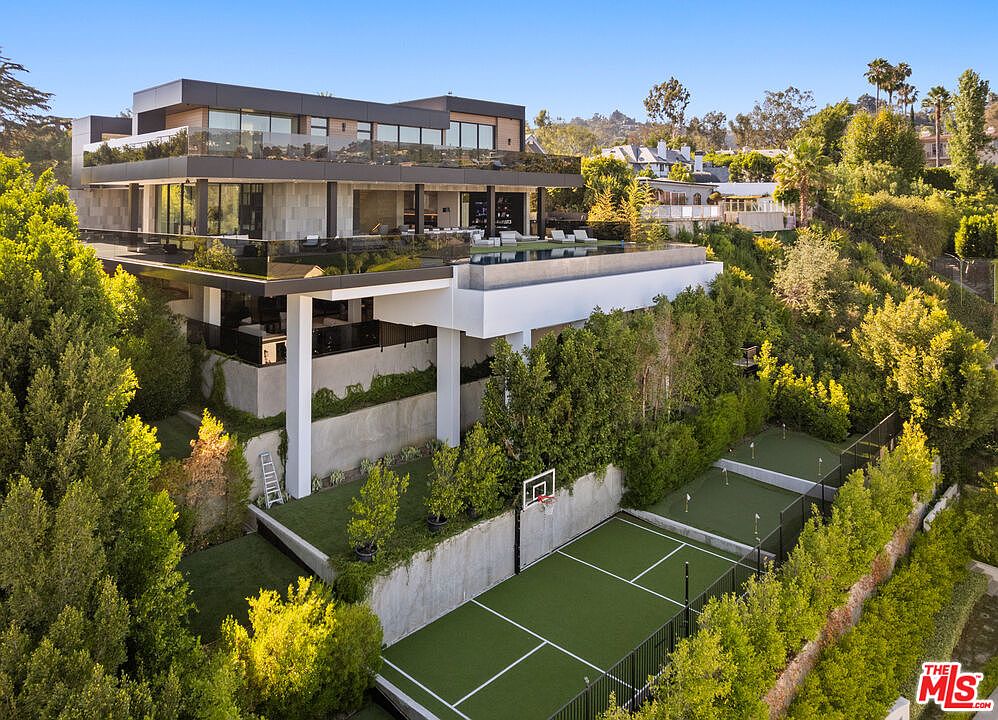 This aerial view showcases a modern luxury home with multiple levels, including a rooftop pool and lounge area. The property features meticulously landscaped grounds with a tennis court, putting green, and lush greenery, creating a private and upscale outdoor environment. The architectural design emphasizes clean lines and expansive glass elements, blending seamlessly with the surrounding landscape.