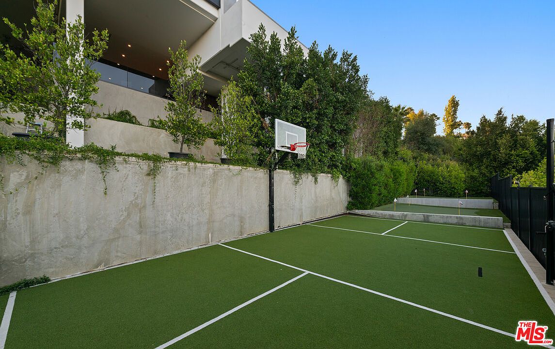 This image showcases a well-maintained outdoor sports area, featuring a basketball hoop and what appears to be a putting green. The area is covered in artificial turf, delineated by white lines, and bordered by a concrete retaining wall and lush greenery. The setting suggests a private and luxurious backyard amenity.