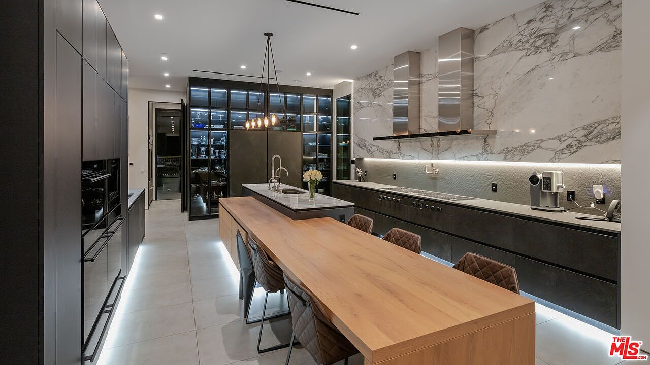 This is a modern kitchen with sleek, dark cabinetry and a large island featuring a light countertop and a wooden extension serving as a dining table. Stainless steel appliances and a marble backsplash add a touch of luxury. The room is well-lit with recessed lighting and pendant lights over the island, creating a sophisticated and functional space.
