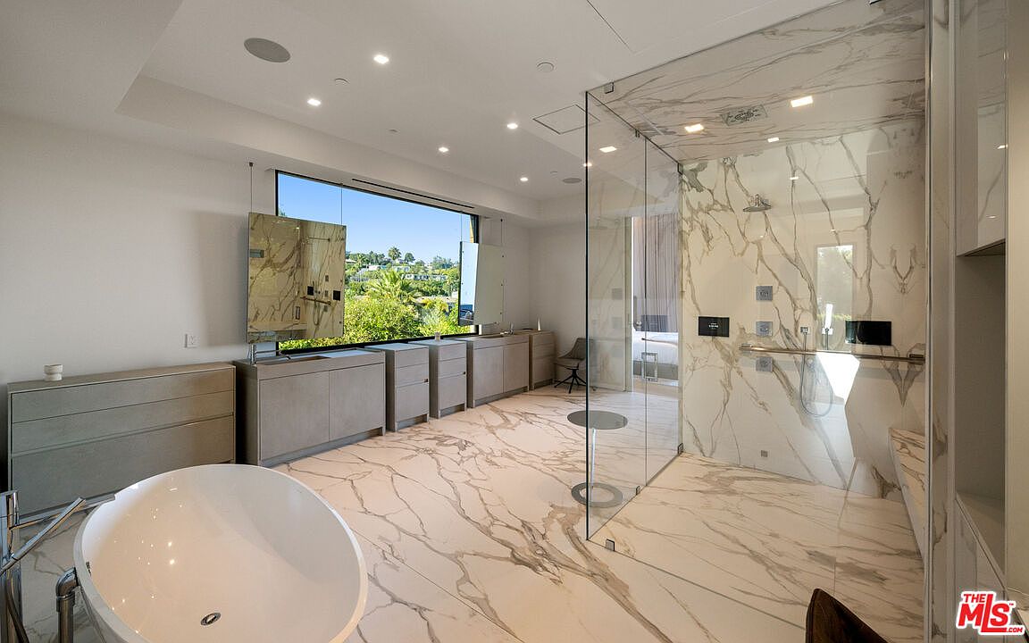 This is a luxurious primary bathroom featuring marble flooring and walls, a freestanding soaking tub, and a glass-enclosed shower. The room has a modern design with clean lines and a neutral color palette. A large window provides natural light and a view of the outdoors, while a vanity with ample storage completes the space.