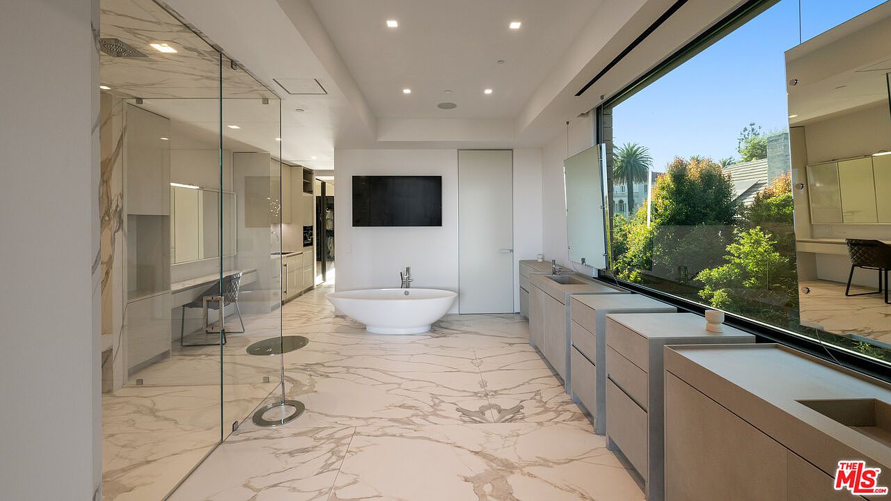 This is a luxurious primary bathroom featuring a freestanding bathtub, a large flat-screen TV, and dual vanities with modern fixtures. The bathroom has marble flooring and a large window offering natural light and views of the outdoors. A glass-enclosed shower area and a separate vanity area are also visible, enhancing the spa-like atmosphere.