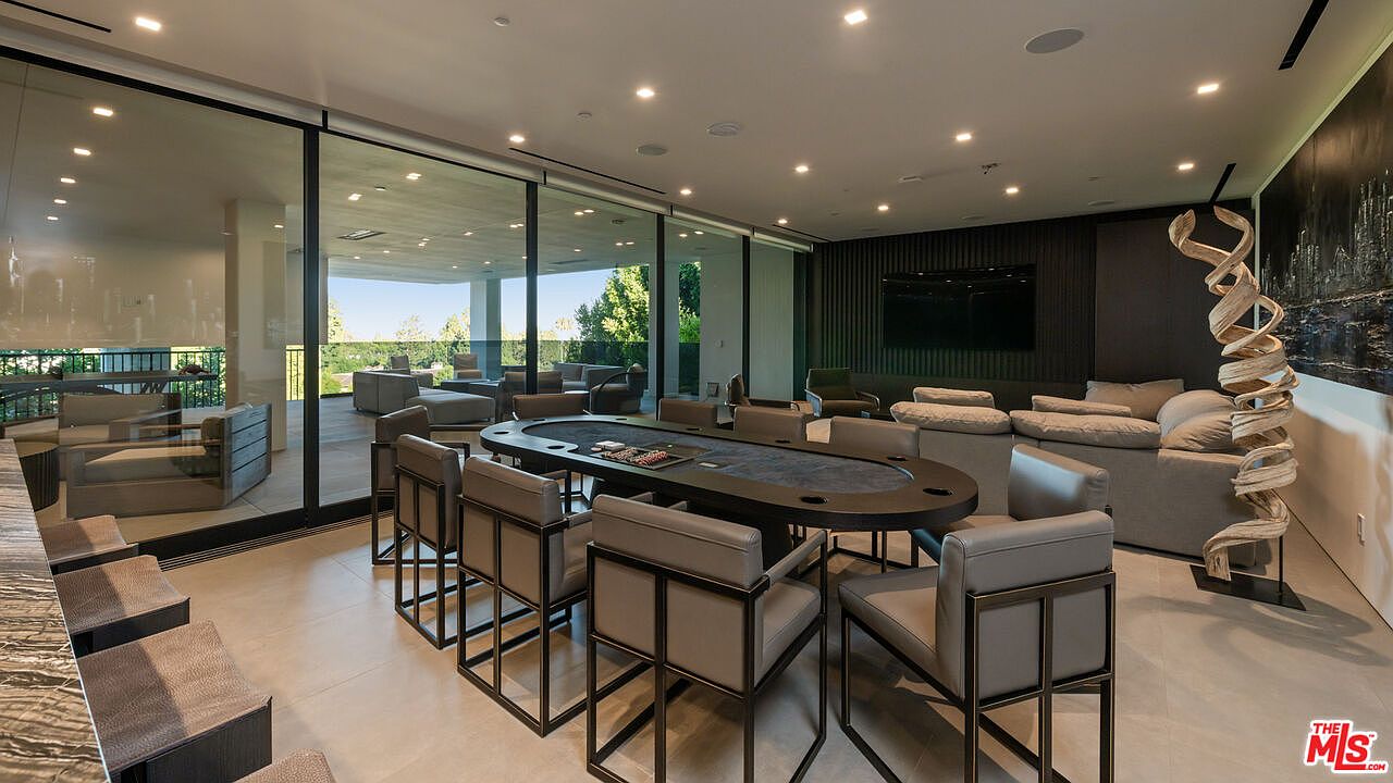 This interior shot showcases a modern living room with a poker table as a central feature, surrounded by stylish chairs. A large sliding glass door provides ample natural light and a view of an outdoor lounge area. The room also includes a comfortable sofa and a unique sculptural art piece, contributing to a sophisticated and inviting atmosphere.