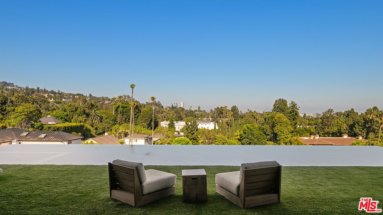 This image showcases a luxurious infinity pool overlooking a lush, green landscape with scattered houses and trees under a clear blue sky. Two modern lounge chairs and a small table are positioned on the manicured lawn in the foreground, inviting relaxation and enjoyment of the scenic view. The overall impression is one of tranquility and high-end living.